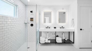 A modern bathroom with white tiled walls, a glass shower enclosure, a white sink with dual mirrors, and a towel rack with neatly folded towels.