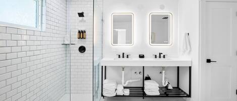 Un baño moderno con paredes de azulejos blancos, cabina de ducha de vidrio, lavamanos blanco con espejos dobles y un perchero con toallas dobladas cuidadosamente.