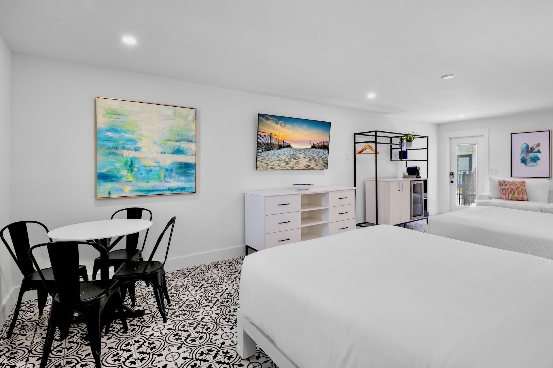 Una habitación de hotel moderna con una cama grande, un rincón de comedor con mesa y sillas, un aparador blanco y dos cuadros en las paredes.