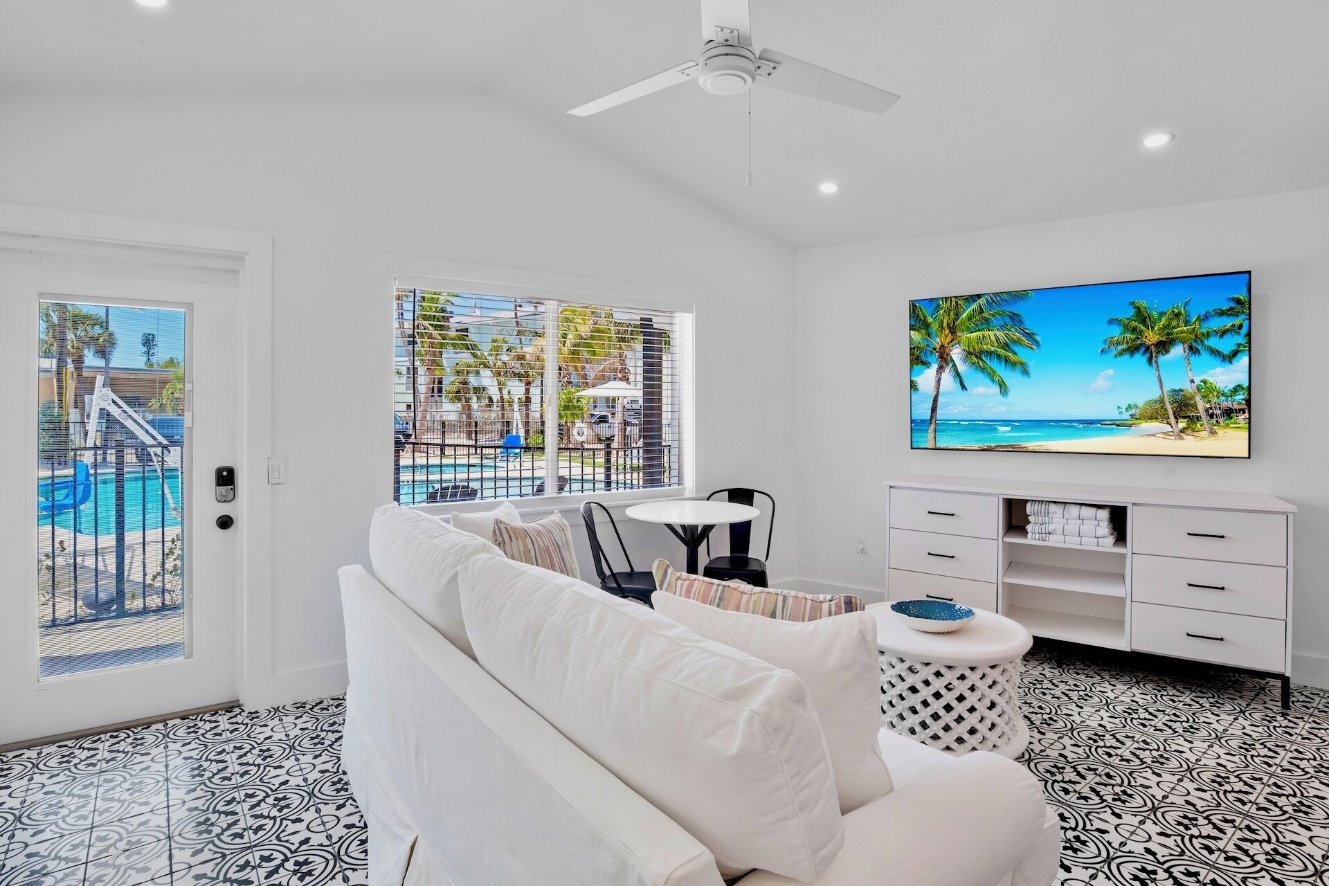 Ein modernes Wohnzimmer mit einem weißen Sofa, einem Fernseher an der Wand und Blick auf einen Pool und Palmen.
