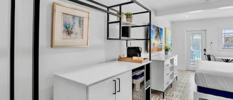 A modern bedroom with a bed, a desk, and a coffee machine. There are framed pictures on the wall and a window with a view of the outside.
