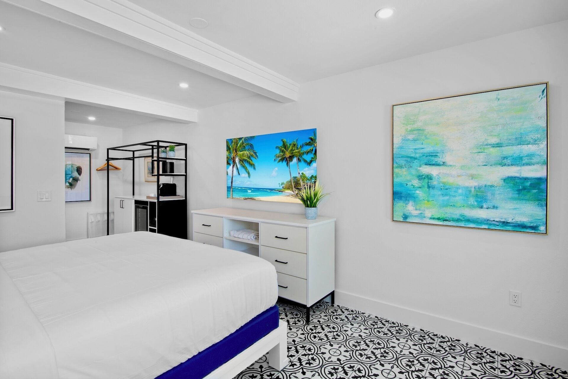 A modern bedroom with a large bed, a white dresser, a desk, and a painting of a beach scene.