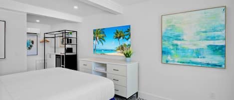 A modern bedroom with a large bed, a white dresser, a desk, and a painting of a beach scene.