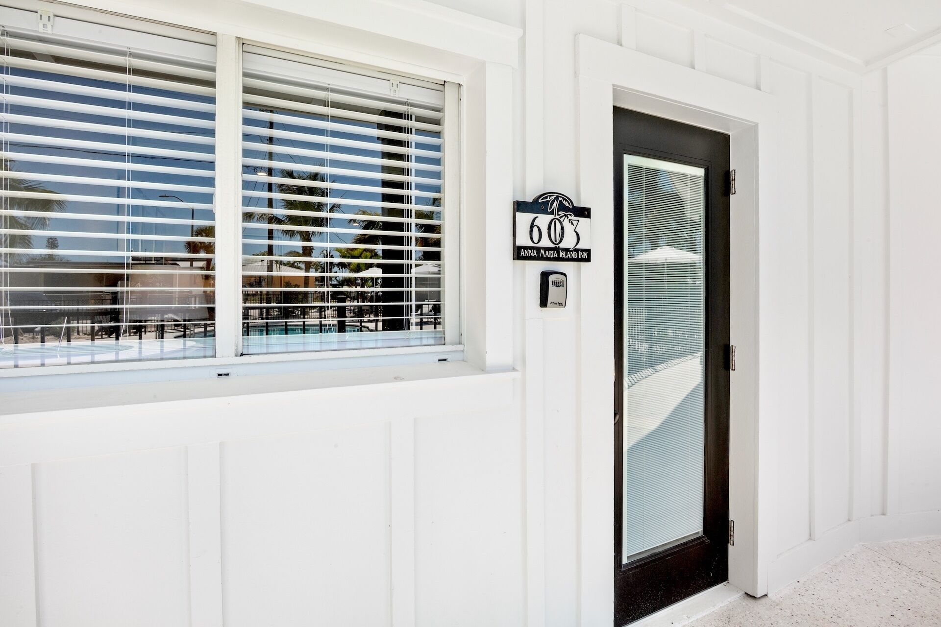 A white building with a black door and a window with horizontal blinds. A sign above the door reads "603 Anna Maria Island Inn."