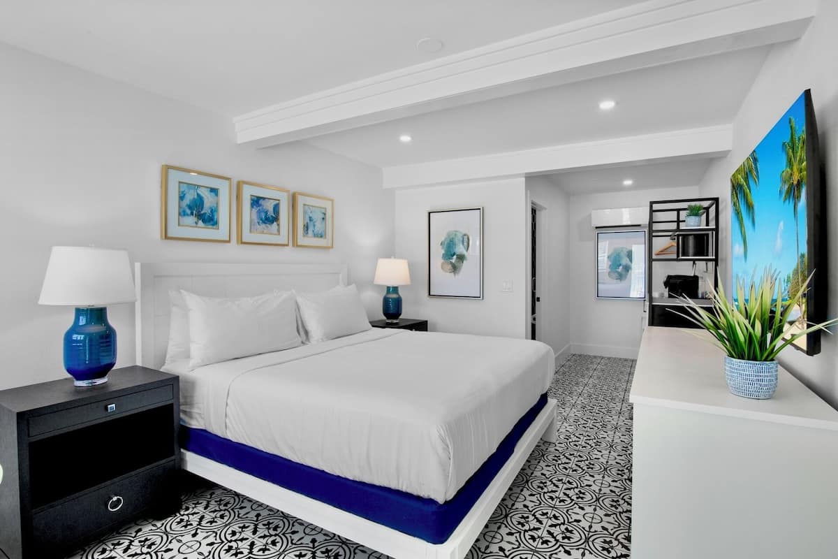 A modern hotel room with a large bed, a desk, and a TV. There are framed artworks on the wall and a plant on the desk.