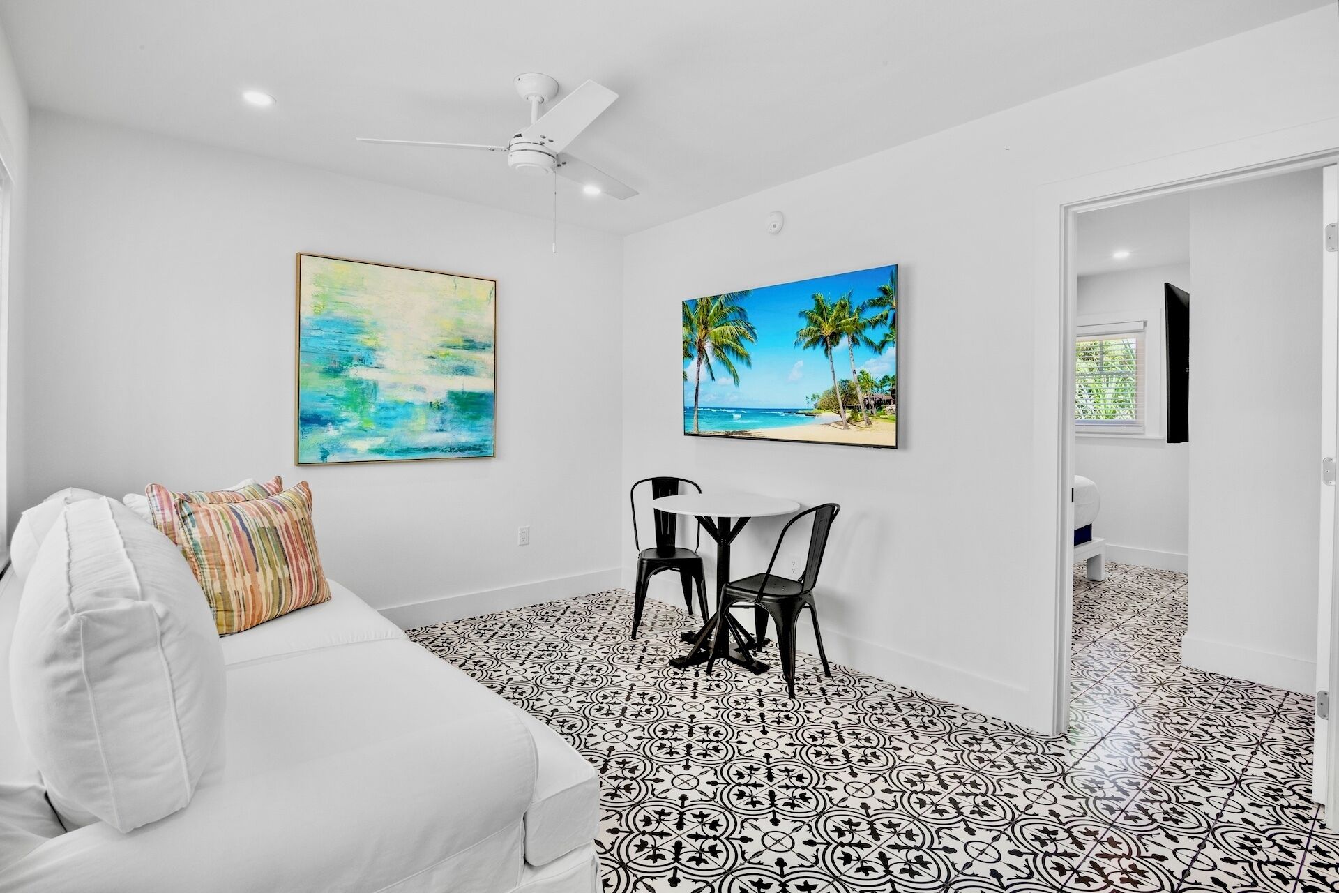 A bedroom with a white bed, a ceiling fan, two paintings, a small table with chairs, and a patterned floor.