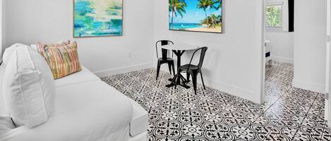 A bedroom with a white bed, a ceiling fan, two paintings, a small table with chairs, and a patterned floor.