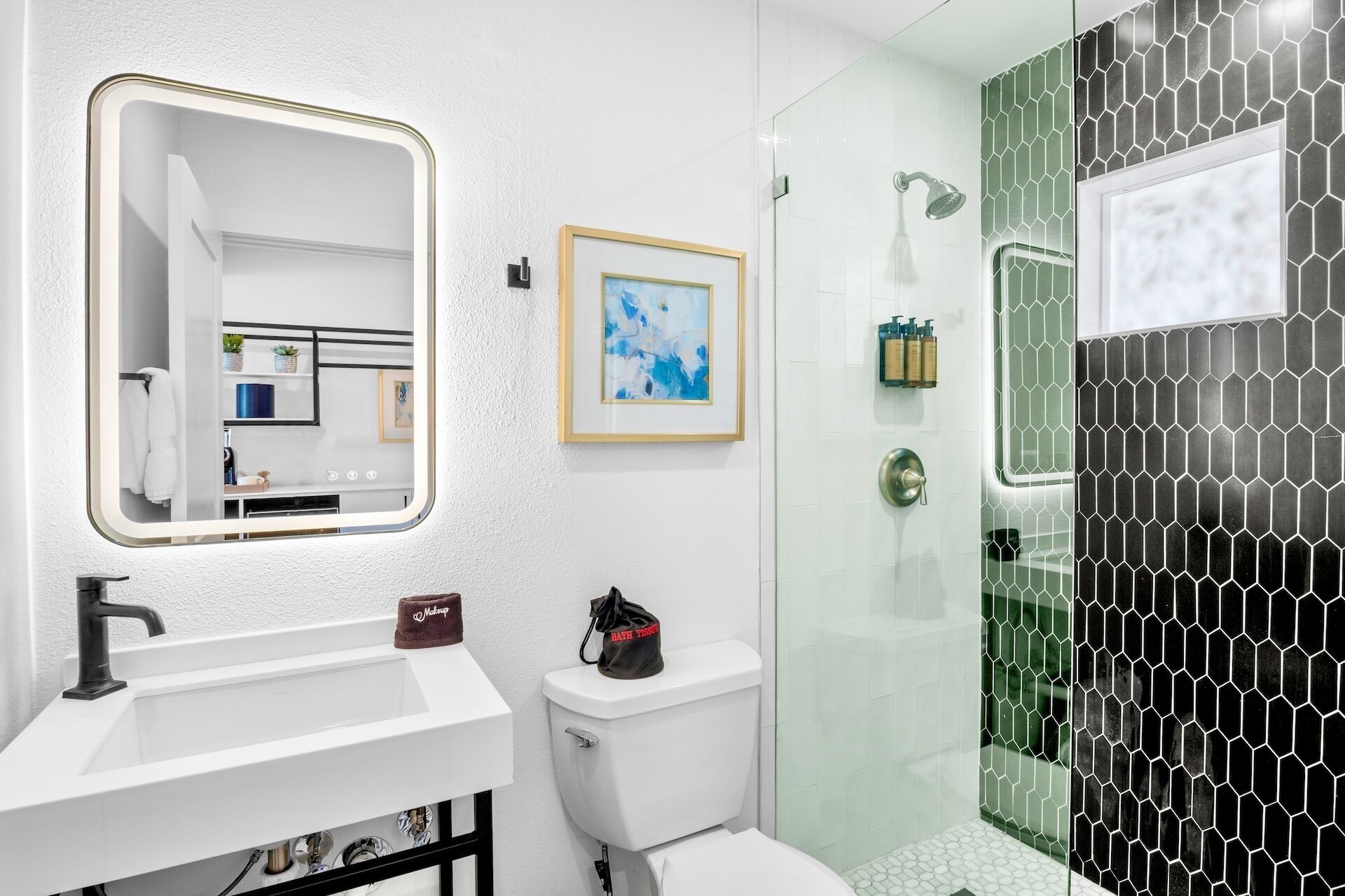 A modern bathroom with a white sink, a framed abstract painting, a glass shower enclosure, and a toilet with a bag labeled "bath tissue".