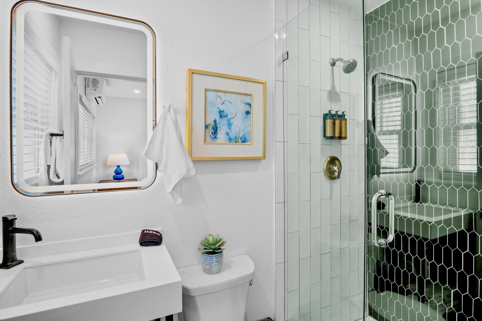 Un baño con un lavabo blanco, un cuadro abstracto en marco, una pequeña planta en maceta y una cabina de ducha de vidrio con un patrón hexagonal.