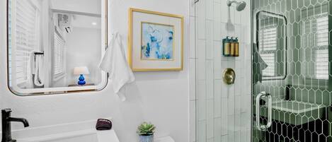 A bathroom with a white sink, a framed abstract painting, a small potted plant, and a glass shower enclosure with a hexagonal pattern.
