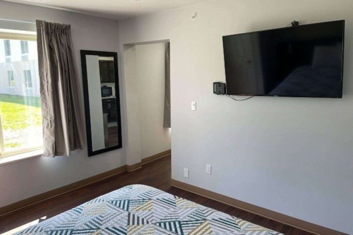 A bedroom with a bed, a TV mounted on the wall, a window with curtains, and a door leading to another room.