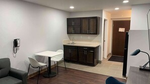 A compact kitchen with dark cabinets, a white countertop, and a small sink. There is a white dining table with two chairs, a grey sofa, and a desk with a lamp. - Studio 6 Peoria, IL (Peoria)