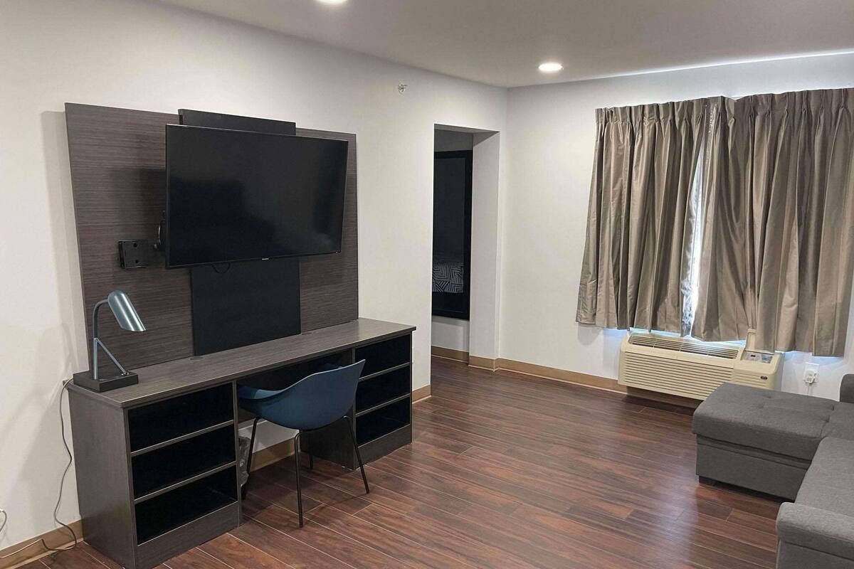 Una sala de estar moderna con un televisor de pantalla plana montado en una pared de madera oscura, un sillón azul, un sofá gris y una ventana con cortinas.