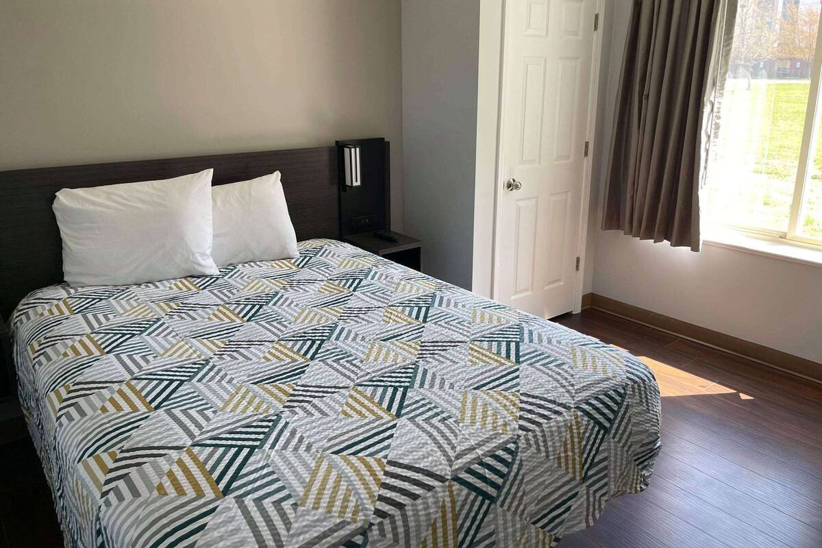 Una cama bien hecha con una colcha estampada, una mesita de noche con un control remoto, una puerta cerrada y una ventana con cortinas.