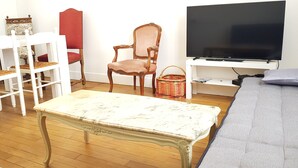A room with a wooden floor, a white table with a marble top, a grey sofa, a red chair, a wooden chair, a TV on a white stand, and a wicker basket. - HOUSE in the center of Fécamp - 90 M2 (FECAMP)