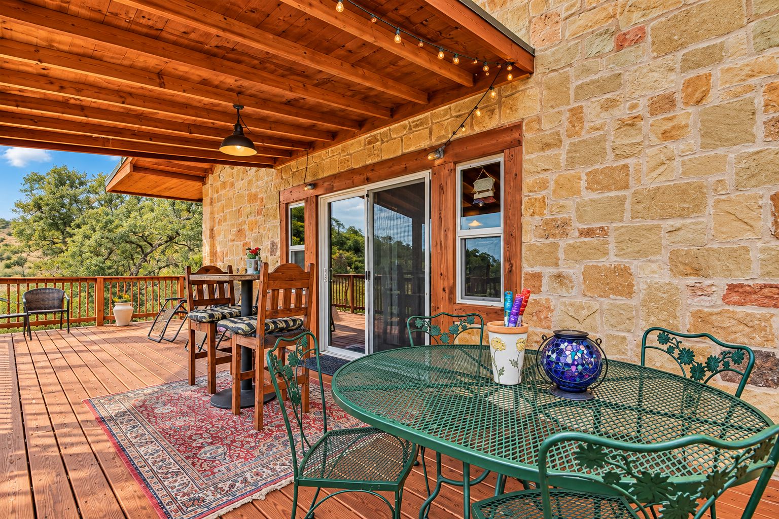 A covered patio with wooden flooring, green metal chairs, a table, and a decorative vase.