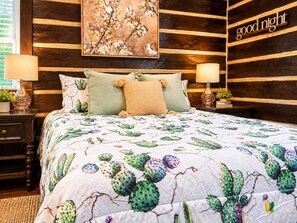 A bedroom with a cactus-themed bedspread, two bedside lamps, a nightstand with a plant, and a wall-mounted sign that reads 'good night'. - Rustic BLUEBONNET CABIN with Views-Near Hidden Falls Park-2 acres, 2 min to lake (Marble Falls)