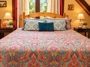 A bed with a patterned bedspread, two pillows, a nightstand with a lamp, and a window with curtains. - Rustic BLUEBONNET CABIN with Views-Near Hidden Falls Park-2 acres, 2 min to lake (Marble Falls)