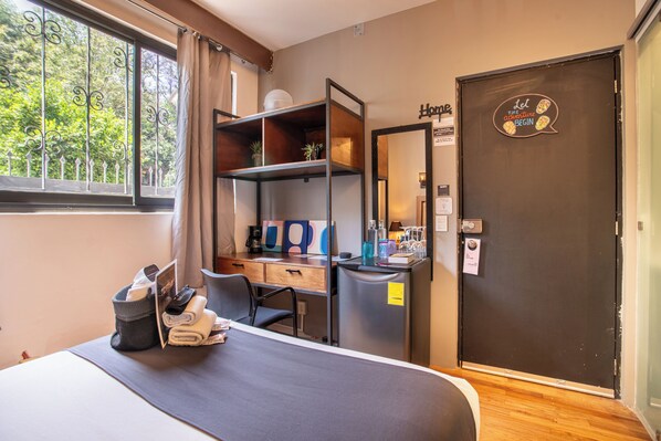 A hotel room with a bed, a desk with a computer, a chair, a small refrigerator, and a door with a sign.