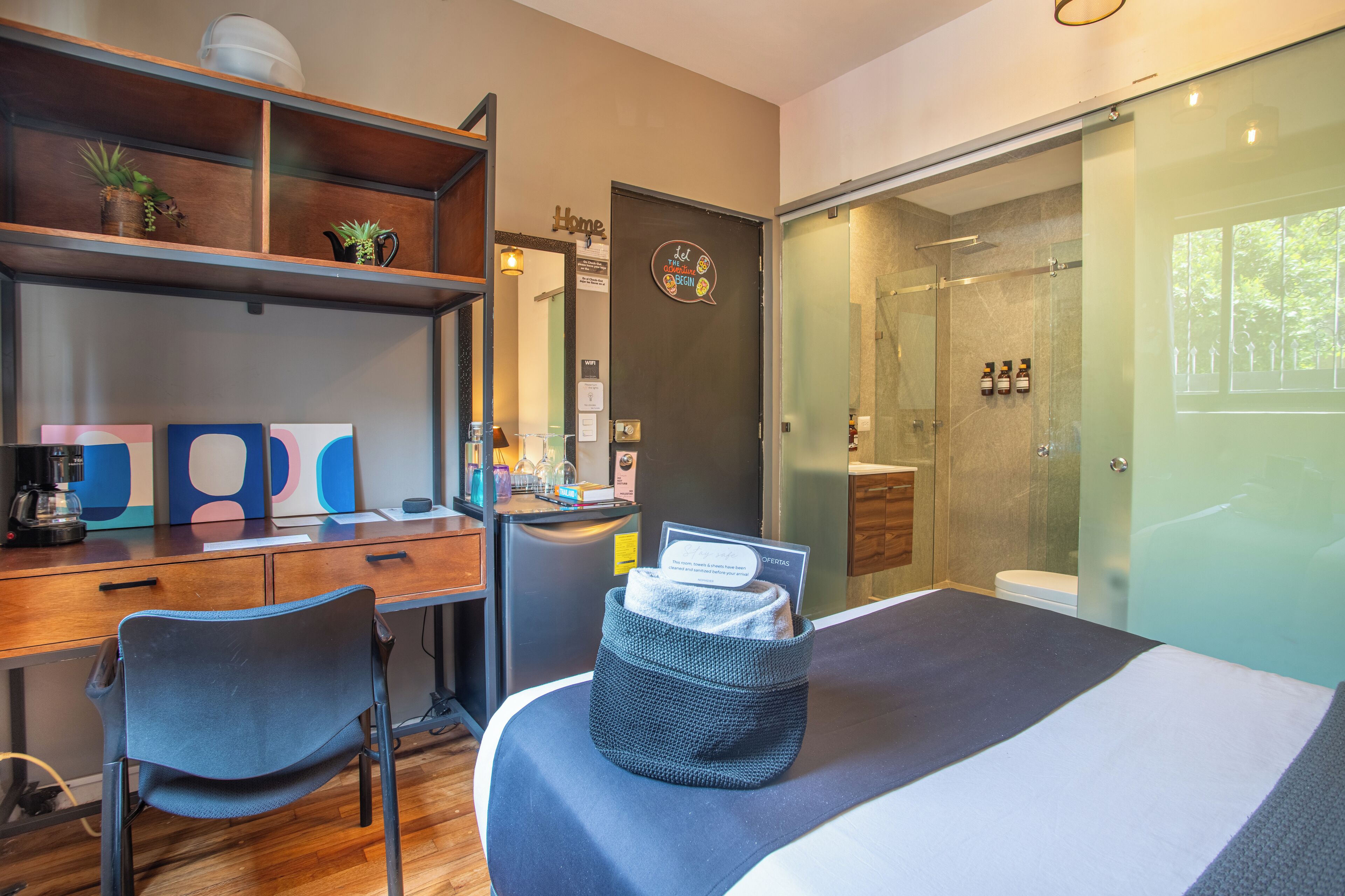 A modern hotel room with a desk, chair, and a bed with a storage basket. There is a shower area with a glass door and a toilet visible.
