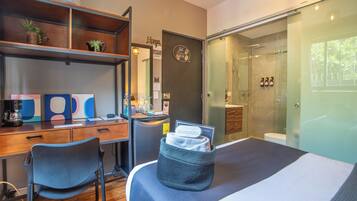 A modern hotel room with a desk, chair, and a bed with a storage basket. There is a shower area with a glass door and a toilet visible.
