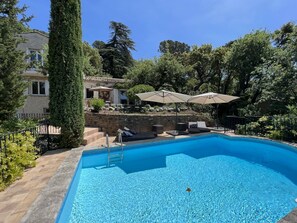 A swimming pool with lounge chairs and umbrellas, surrounded by trees and a stone wall.