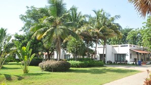 A white building with a tiled roof, surrounded by lush greenery and palm trees. - Thappa Gardens Resort (Karaikudi)