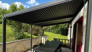 Eine Außenpatio mit überdachtem Tisch und Stühlen, einem steinbelagten Boden und Blick auf die umliegende Vegetation.