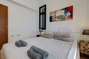 Un lit bien fait, agrémenté d’oreillers et de serviettes roulées, une table de chevet avec une lampe, et un tableau abstrait aux couleurs vives accroché au mur.
