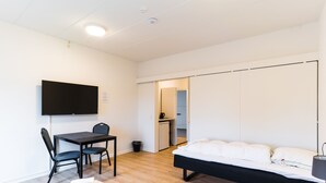 A modern hotel room with a bed, a desk with chairs, a television, and a kitchenette in the background.