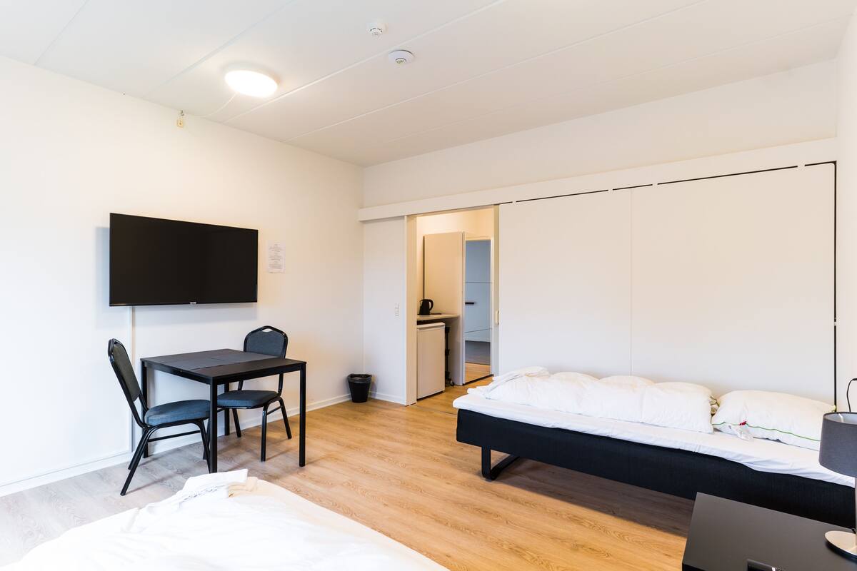 Une chambre d’hôtel moderne avec un lit, un bureau avec des chaises, une télévision et, au fond, une kitchenette.