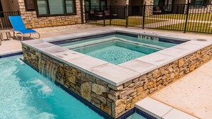 Una bañera de hidromasaje rectangular con paredes de piedra y borde de azulejos, rodeada por una piscina.
