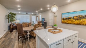Cocina amplia con una isla de mármol, mesa de comedor de madera y sillas. Se observa un cuadro en la pared y un ventilador de techo.