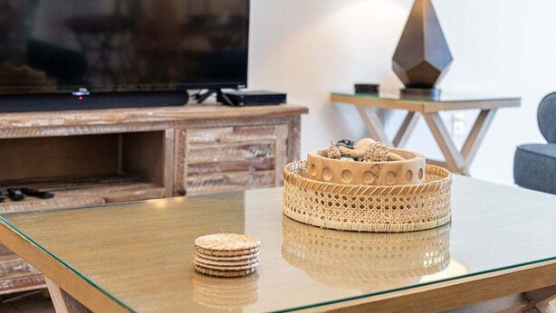 Una sala de estar moderna con una mesa de centro de vidrio, un cuenco decorativo tejido y un televisor sobre un soporte de madera.