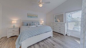 Un dormitorio con cama, mesitas de noche, cómoda, ventilador de techo y ventana con persianas.