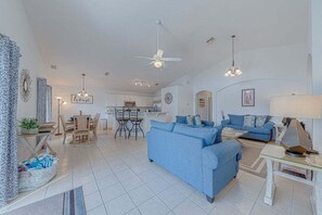 Un vaste salon avec un canapé bleu, un coin repas avec une table et des chaises, une cuisine à l’arrière-plan et un ventilateur de plafond.