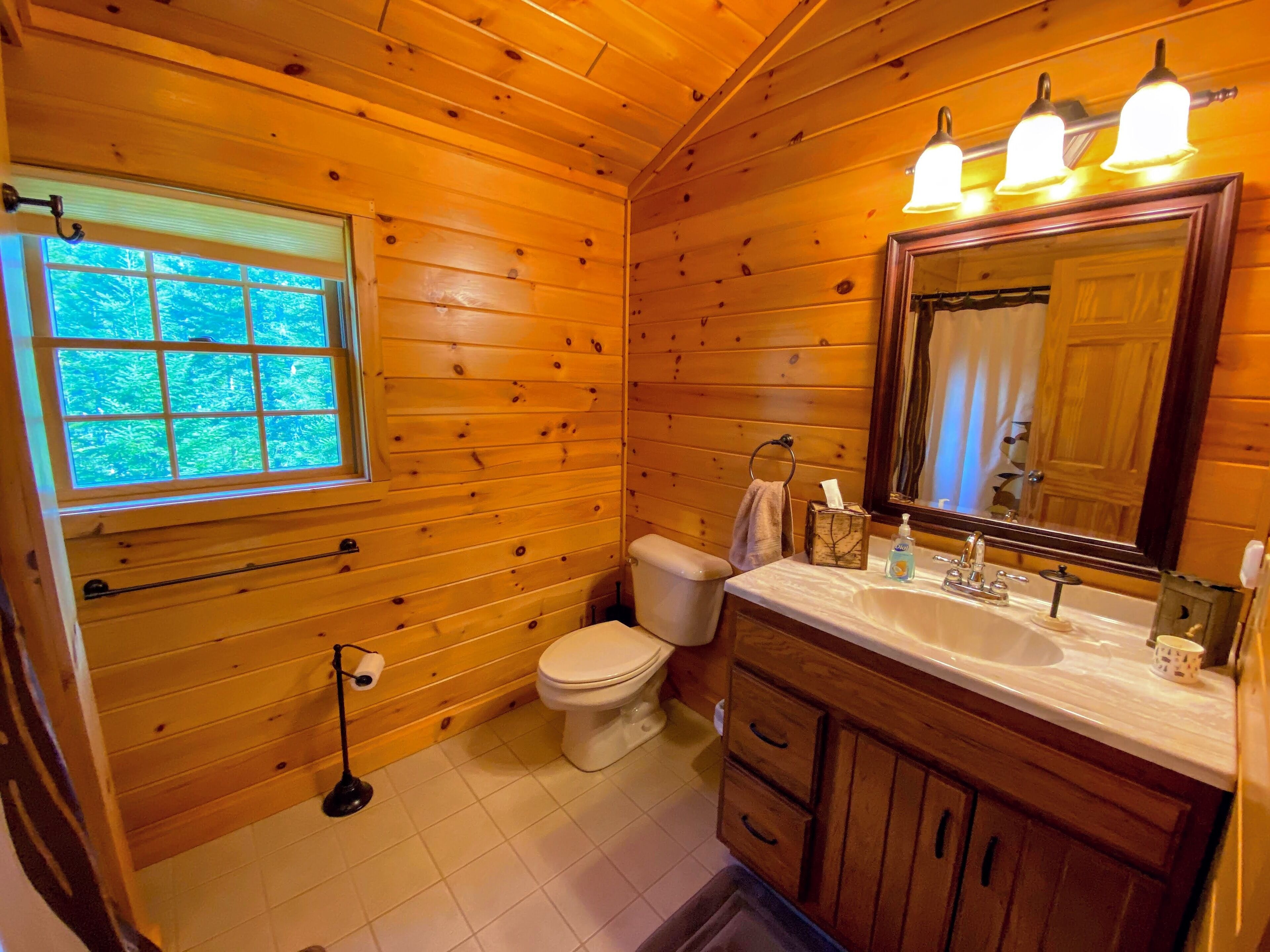 A wooden cabin bathroom with a window, toilet, sink, and mirror.