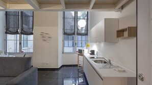 Une cuisine compacte équipée d’un évier, d’un four à micro-ondes et d’un petit plan de travail. On y trouve un canapé gris et une fenêtre avec des rideaux.