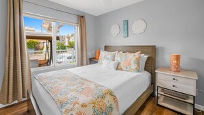 A bedroom with a large bed, a nightstand with a lamp, and a view of the outdoors through a sliding door. - Beach-2-Bay 1br | Mission Beach | AC | Shops (San Diego)