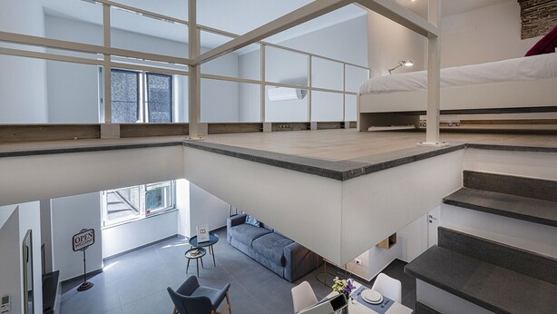 Un ampli espai loft modern amb sostre alt, grans finestres, un llit en alt, zona de menjador i escala.