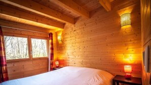 A wooden cabin room with a bed, two bedside lamps, and a window with curtains. - Safari.... (Le Vigen)