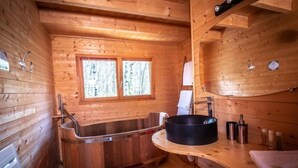 A wooden cabin interior with a wooden bathtub, a sink, and a window with a view of trees. - Safari.... (Le Vigen)