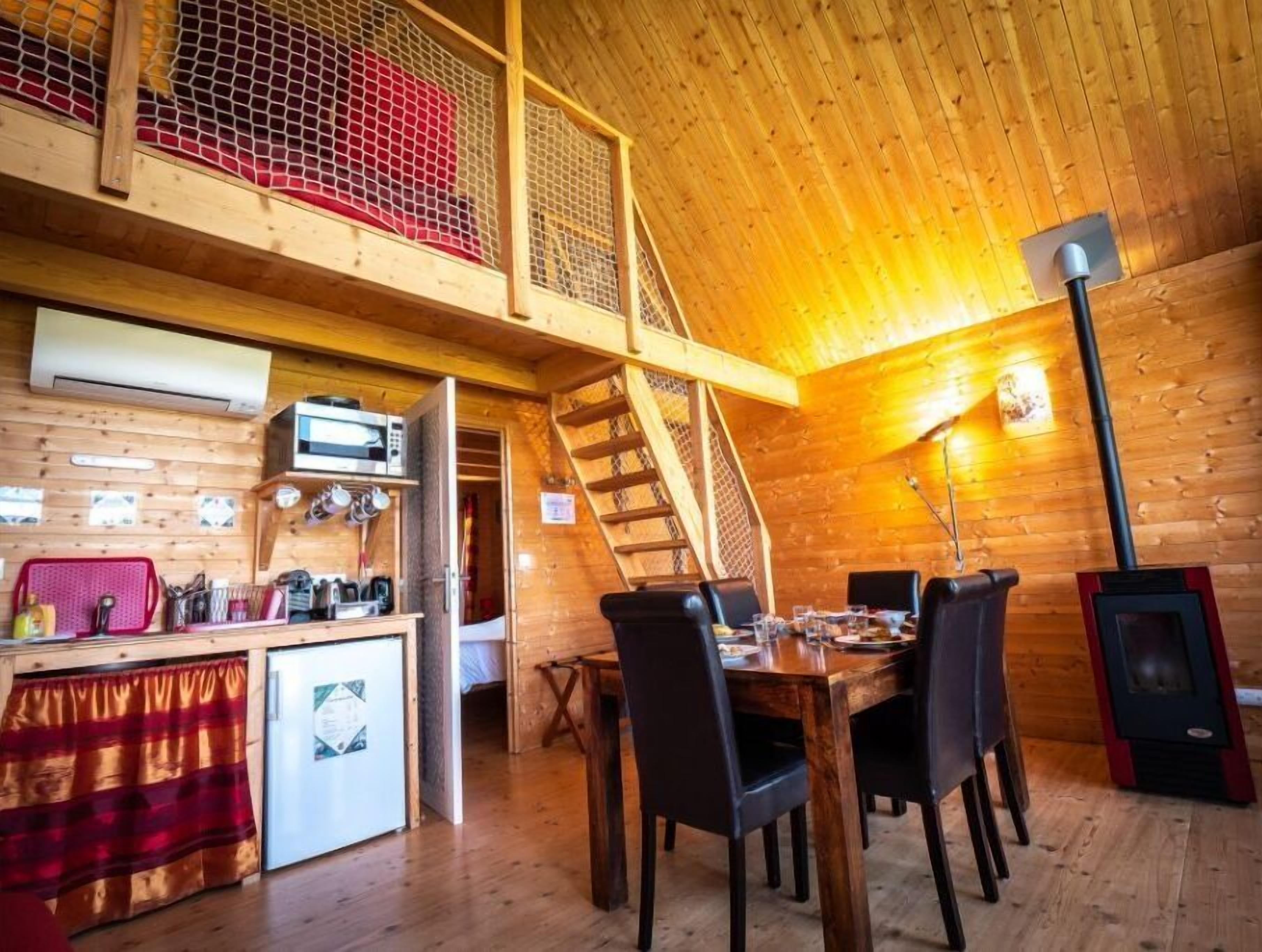 Un chalet confortable avec un intérieur en bois, un coin repas avec des chaises, une petite cuisine équipée d’un réfrigérateur et un escalier menant à une mezzanine.