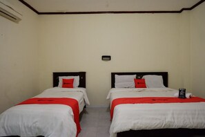 Habitación de hotel con dos camas individuales, cada una con una colcha blanca y roja, y una mesita de noche pequeña con un control remoto.