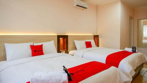 Una habitación de hotel con dos camas, cada una con ropa blanca y detalles rojos, un cabecero de madera y un equipo de aire acondicionado en la pared.