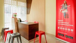 Zona de recepción con un mostrador de madera, taburetes rojos, un gran cartel de RedDoorz y una ventana con persianas.