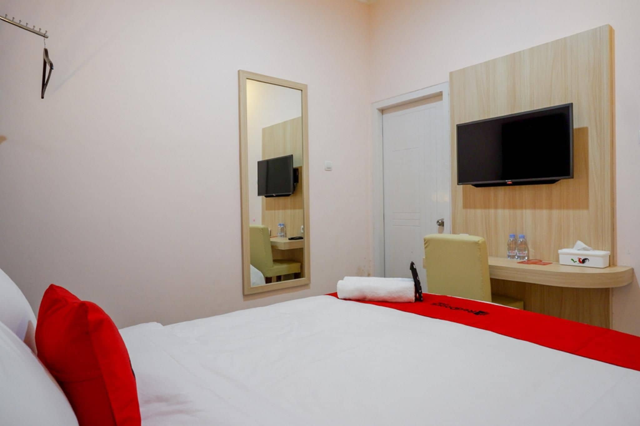 A hotel room with a bed, a TV mounted on the wall, a mirror, a desk with a chair, and a red accent wall.