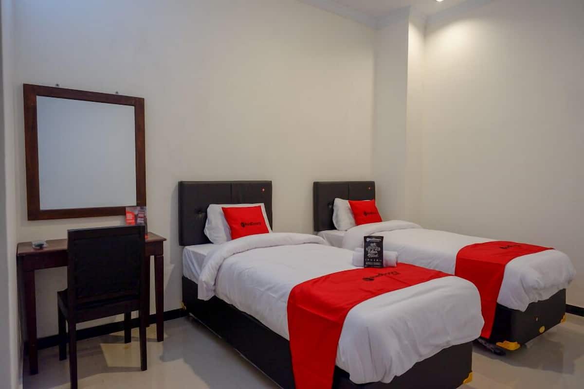 A hotel room with two beds, each with a red blanket and white bedding. There is a desk with a chair and a mirror on the wall.