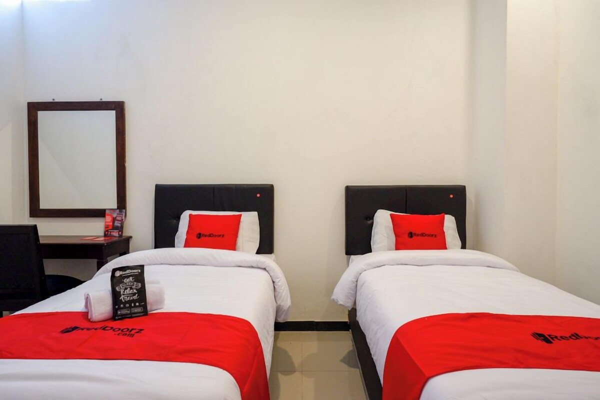 Ein Hotelzimmer mit zwei Einzelbetten, jeweils mit einer rot-weißen Tagesdecke und passenden Kissen. Es gibt einen Schreibtisch mit einem Stuhl und einen Spiegel an der Wand.