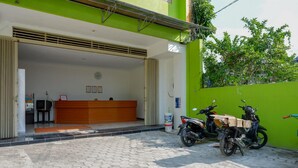 A reception area with a wooden desk, a clock on the wall, and a green wall. There are two motorcycles parked outside. - Reddoorz @ Jalan Wonosari Km.9 (Yogyakarta)